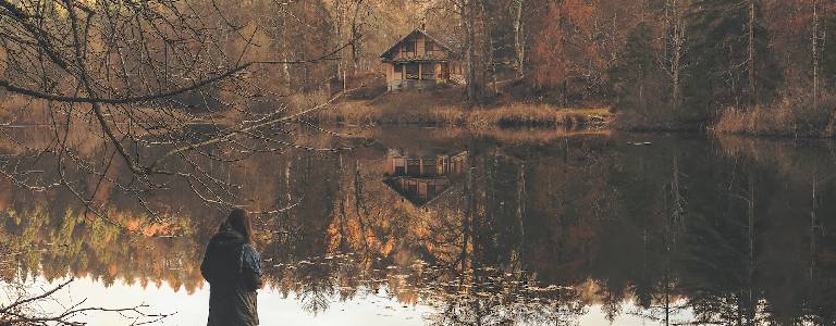 A cabin on the lake surrounded by autumnal trees and a woman looking on