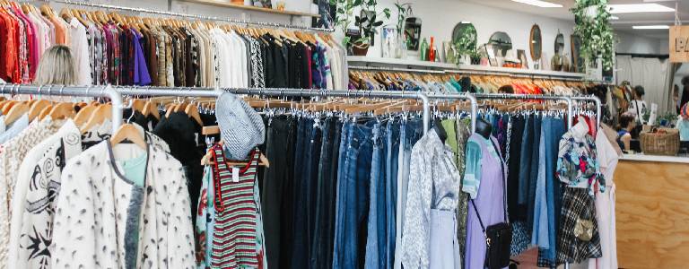 Thrift store clothing racks with woman browsing in the background.