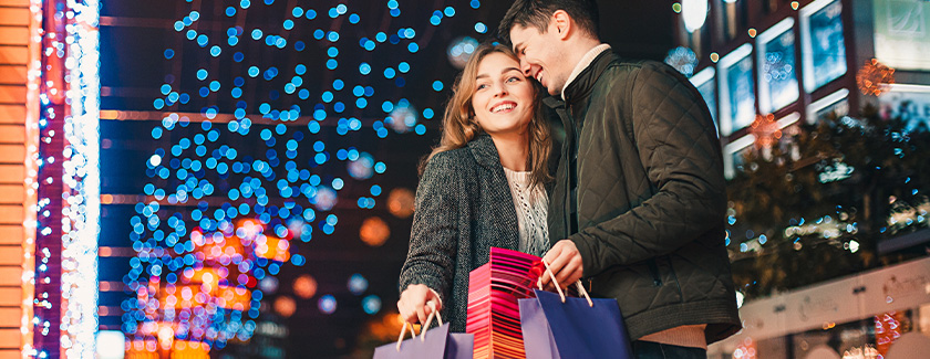 A man and woman smiling outside holding onto shopping bags with Christmas lights sparkling in the background