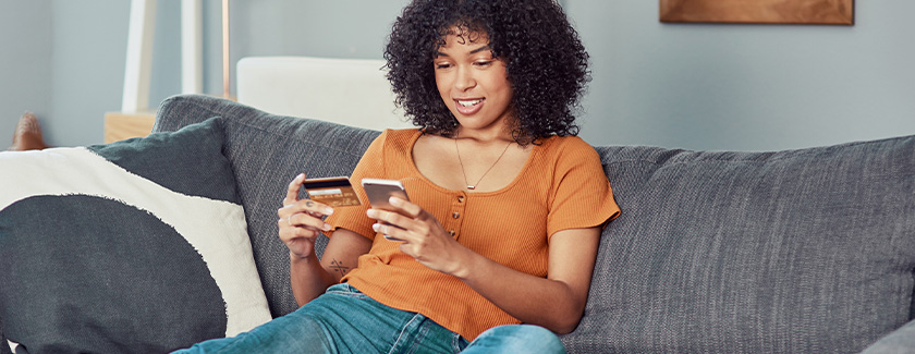 Woman sitting on the couch, holding cellphone in one hand and credit card in another.